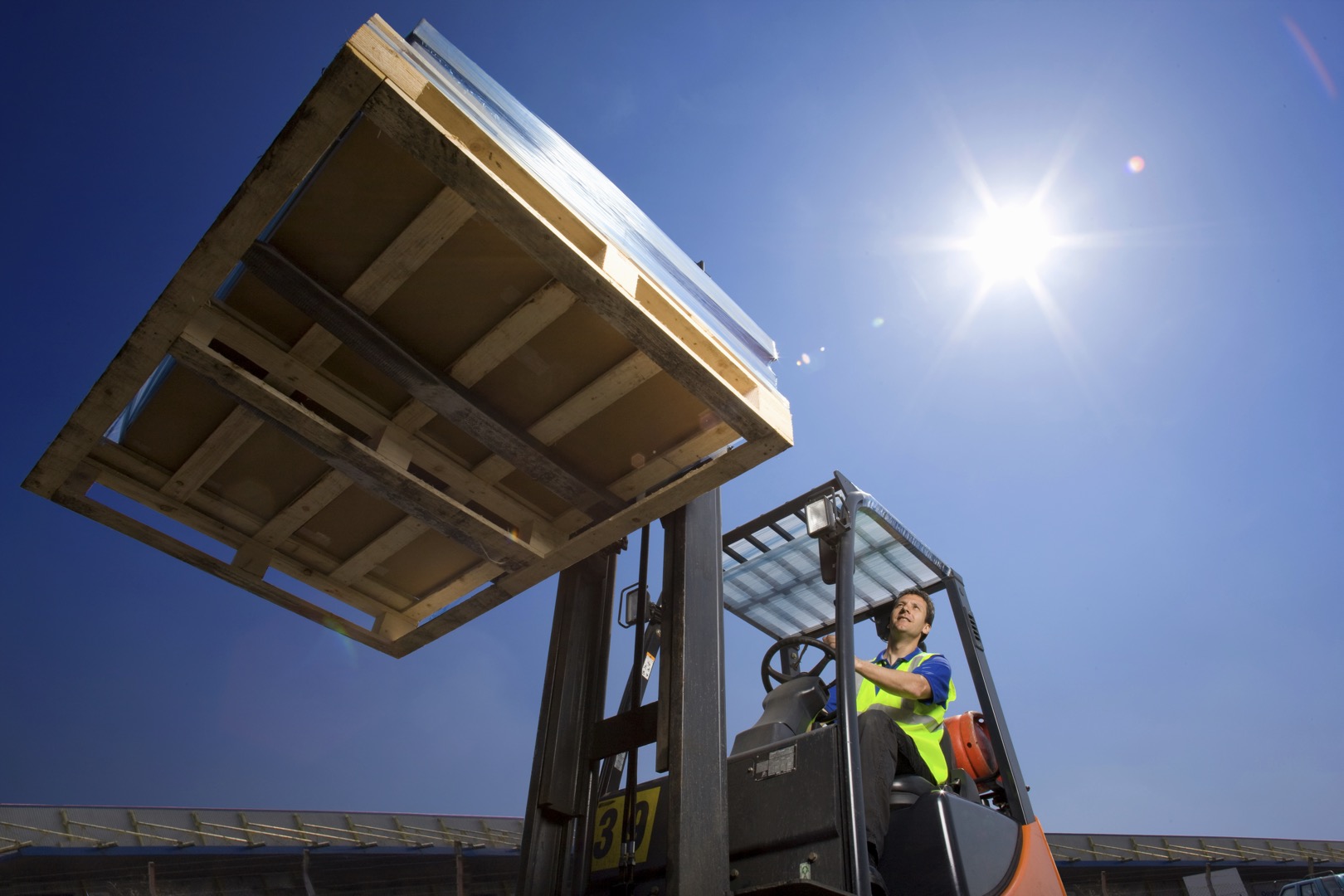 A wide low angle shot of a worker in safety vest using forklift to move the merchandise on a sunny day with a lens flare; Shutterstock ID 1840648450; purchase_order: -; job: -; client: -; other: -